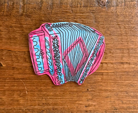 A hand-drawn or painted accordion-shaped item with pink and light blue sections outlined in black, placed on a wooden surface.