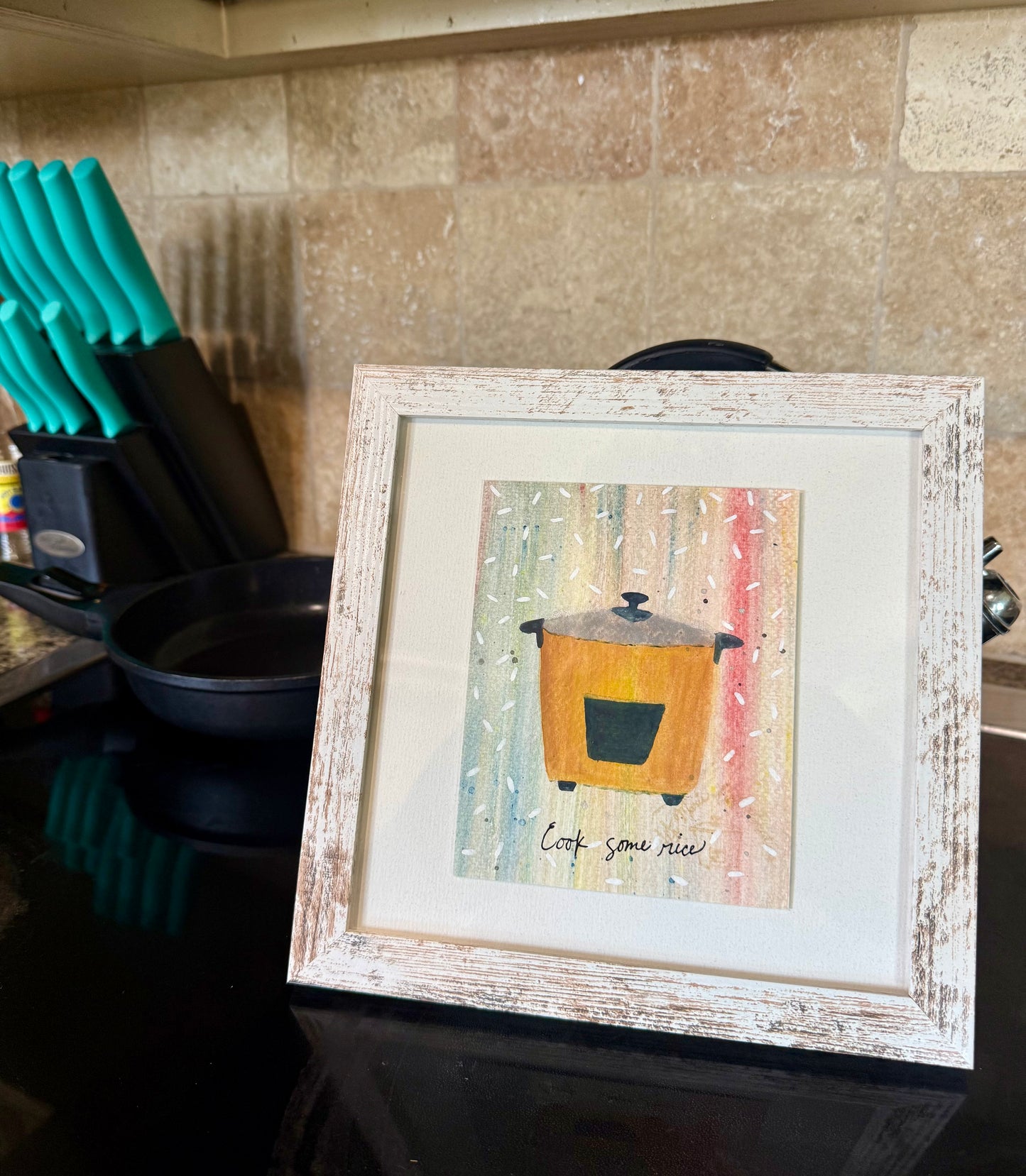 Framed artwork of a ricepot on a kitchen counter with knives and pots in the background.