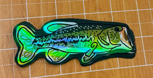 Large mouth bass sticker with an iridescent holographic finish, in green, displayed on a wooden cutting mat.