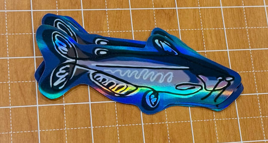 Catfish stickerr with an iridescent holographic finish, in blue, displayed on a wooden cutting mat.